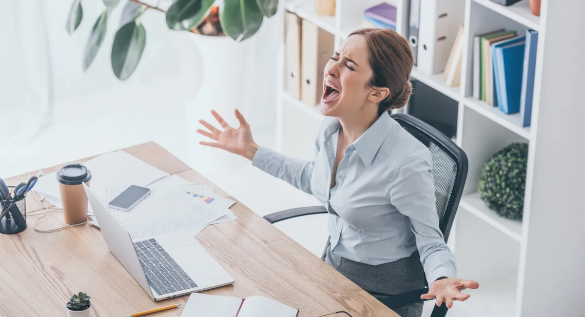 Stress auf der Arbeit: Frustrierte Frau im Office-Look sitzt im Büro vor ihrem Schreibtisch und schreit