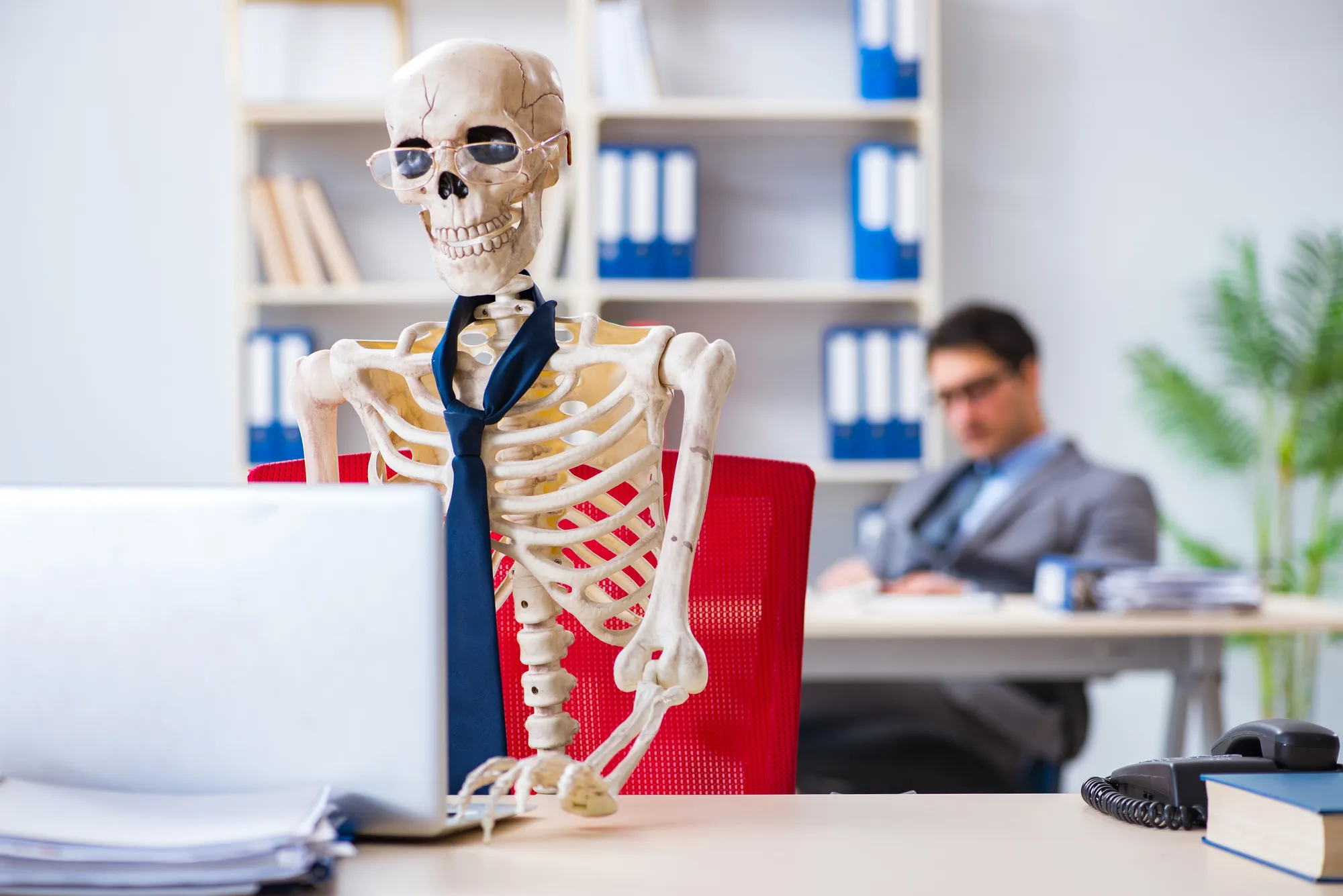 Innerliche Kündigung: Ein Skelett mit Krawatte sitzt im Vordgrund am Arbeitsplatz. Im Hintergrund sitzt ein Mann im Office-Look.