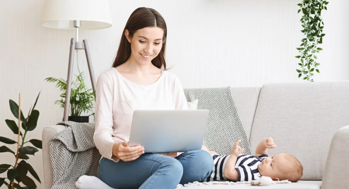 Beruflicher Neustart nach Elternzeit: Sitzende Frau auf dem Sofa mit Laptop. Auf dem Sofa liegt das kleine Kind.