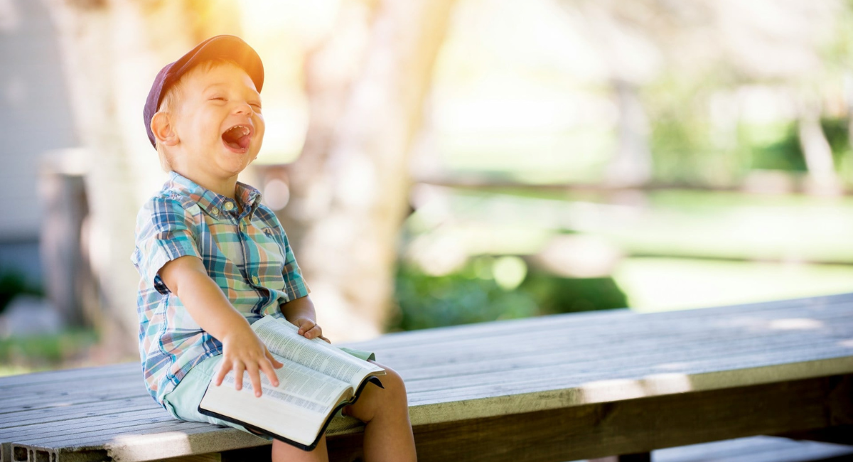 Resilienz bei Kindern: Lachendes Kind mit aufgeschlagenen Buch auf einer Sitzbank bei gutem Wetter